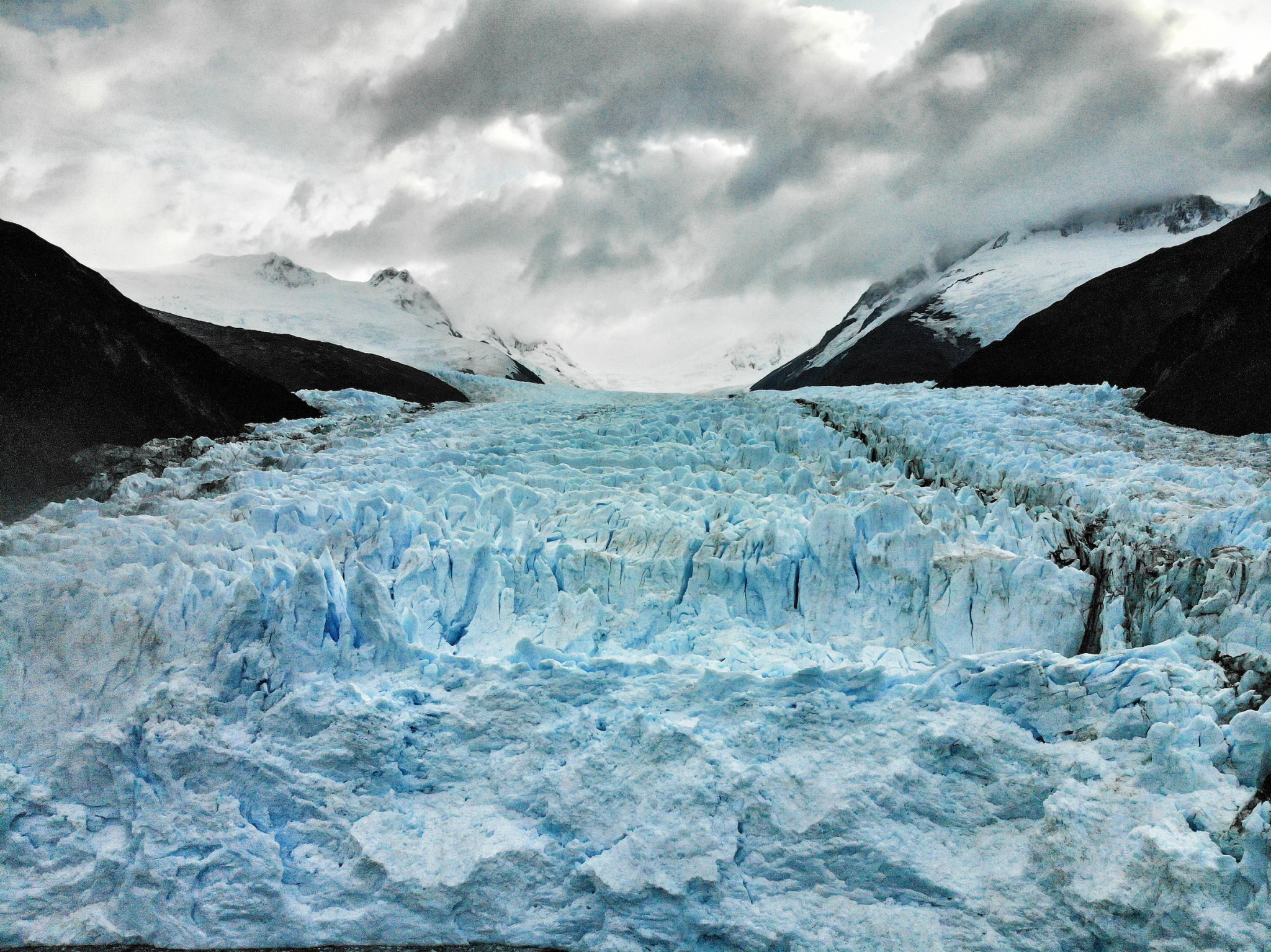 Expansive aerial view of a blue glacier descending between dark mountain slopes beneath heavy clouds.
