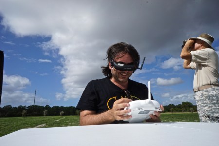 Photographer operating an early drone controller while wearing Fat Shark FPV goggles in an open field under a cloudy sky.