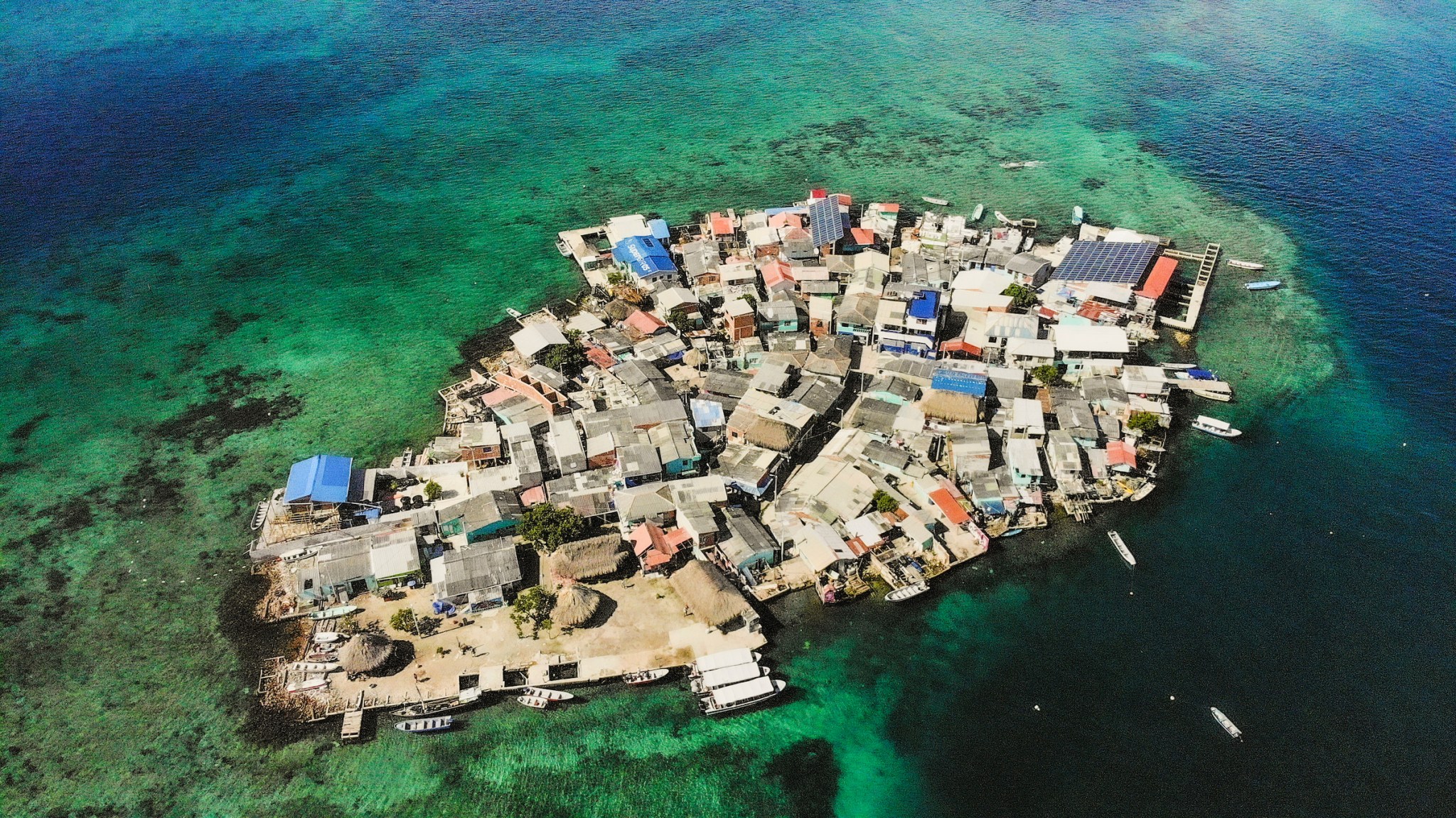 Drone photograph of a densely populated small island surrounded by turquoise water, showing houses, rooftops, boats, and the surrounding sea.